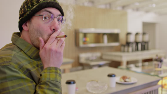 Man in a plaid shirt and beanie smokes a rolling paper cigarette in a modern café