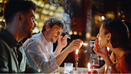 Group using rolling papers to prepare cigarettes, gathered around a candle-lit table in a dim, lively setting.